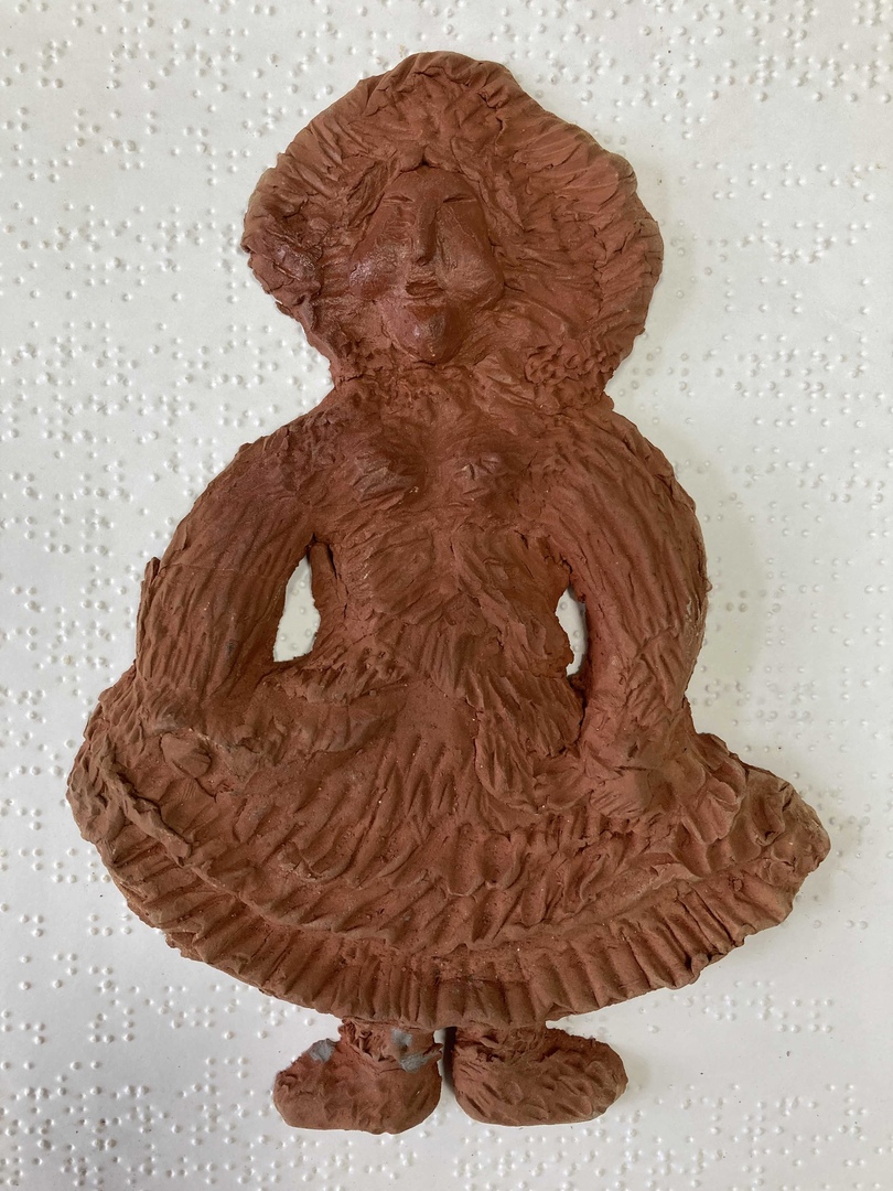 Керамика (2014 г) Работа покрыта коричневой глазурью. На ней изображена женщина-негритянка, От её талии спускается широкая юбка. Её нижний край украшен орнаментом, руки слегка согнуты и упираются в бока в районе талии ( вся из себя купчиха) На голове небольшой чепчик, густые , тёмные, вьющиеся волосы лицу передают определённую пышность. На ногах небольшие ботинки.